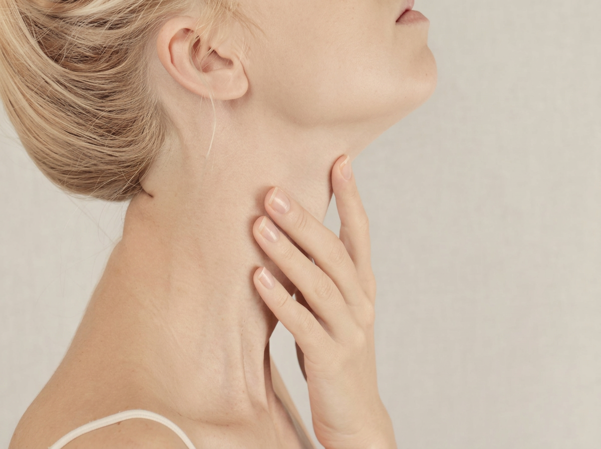 A woman gently holding the front of her neck in soft warm lighting, photographed in a calm beige editorial style