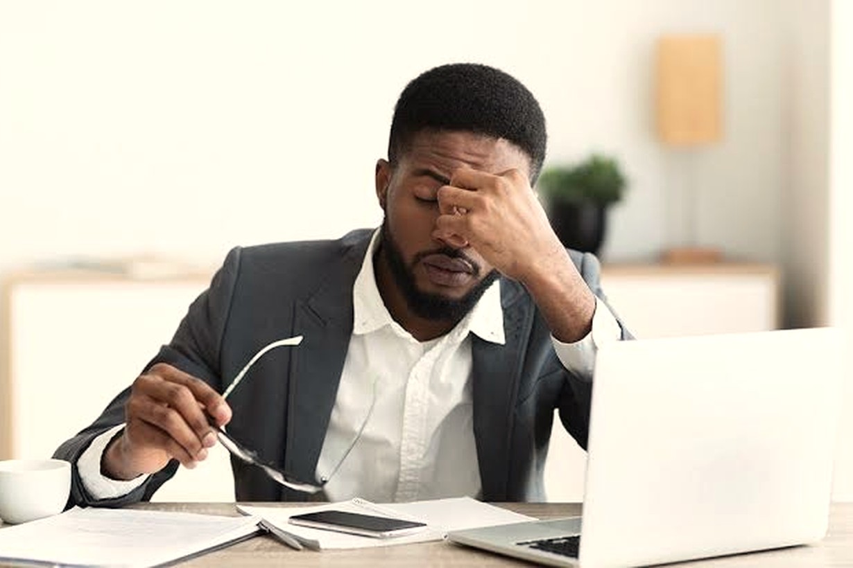 Man in a suit seated at a desk holding his glasses, appearing fatigued and overwhelmed while working